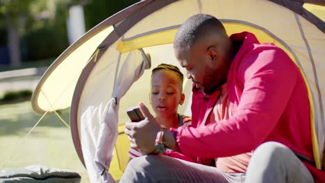 Happy-african-american-father-and-son-in-tent-using-smartphone-in-sunny-garden,-in-slow-motion