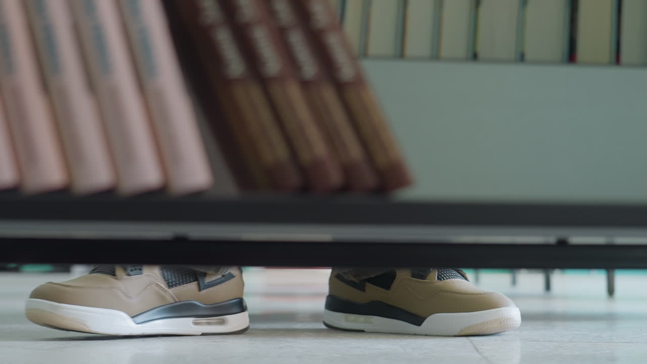 Close up low angle view of person wearing beige sneakers standing on library floor, partially visible under bookshelf with rows of arranged books