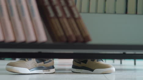 close up low angle view of person wearing beige sneakers standing on library floor, partially visible under bookshelf with rows of arranged books