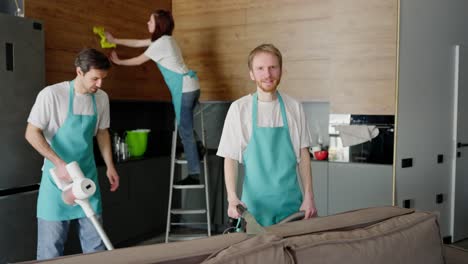 zoom out retrato de un tipo confiado un limpiador rubio en una camiseta blanca y un delantal azul que está limpiando el sofá con una aspiradora junto con sus colegas limpiando la cocina por llamada de una compañía de limpieza