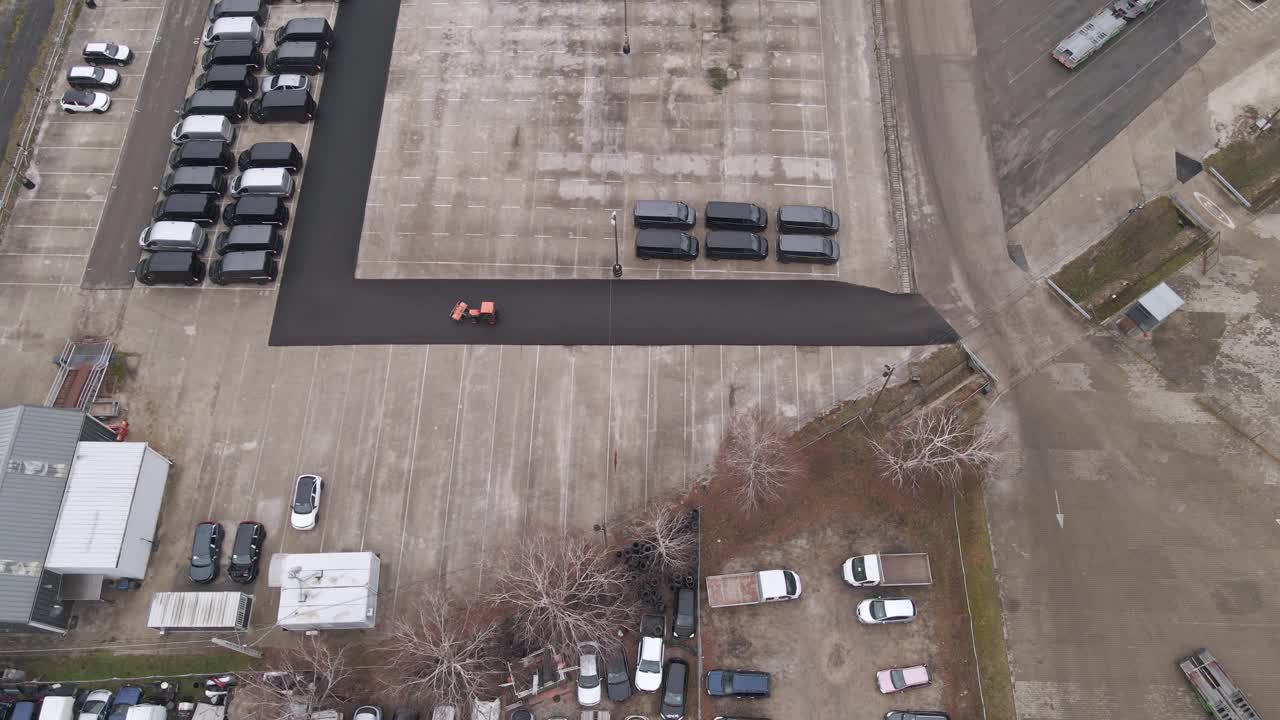 An aerial view of a large parking lot with multiple parked vehicles, including black vans, white cars, and a red tractor driving along a paved road, and a truck leaving the parking lot.