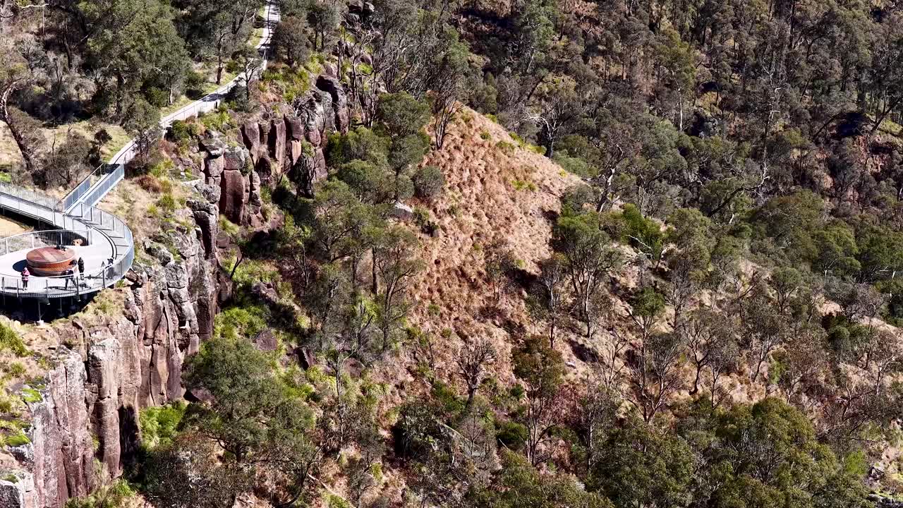 Aerial camera smoothly pans along a scenic clifftop walkway and viewing platform surrounded by forest and rugged terrain in bright daylight