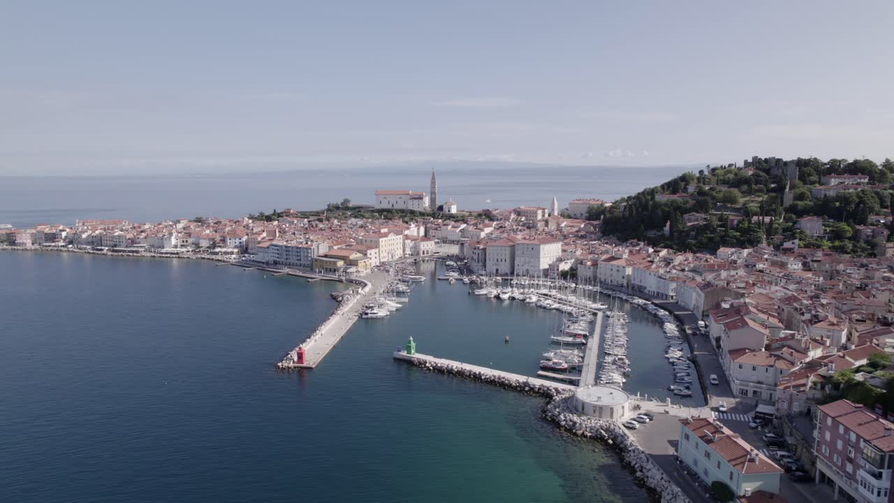 Video with a flat back drone on the coast of Piran in Slovenia, shows the church and the port, discovering the promenade that gives access to the city, you can see the circulation of vehicles