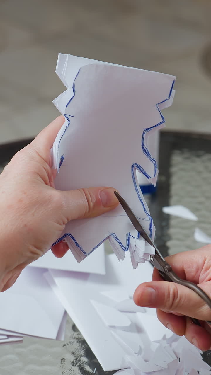 Caucasian hands cutting paper snowflake shapes, closeup on glass tabletop with scissors and glue stick, patient hobbyist trimming layered silhouette for seasonal home decor, soft daylight and cozy