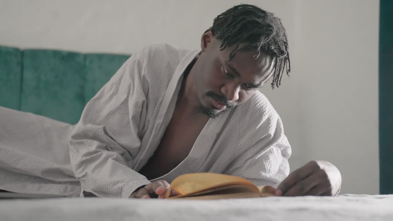 Man in white robe lying on bed, deeply focused on reading large book, hand turning pages slowly. Soft light from window creates peaceful, contemplative atmosphere in calm indoor setting