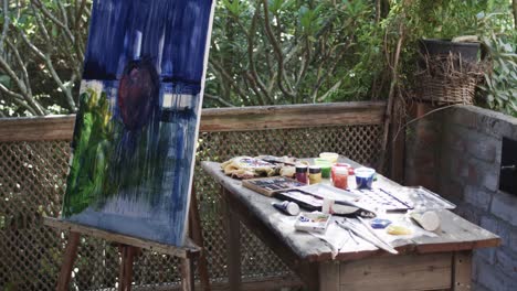 Painting-on-easel-and-paints-and-brushes-on-table-in-sunny-garden,-slow-motion
