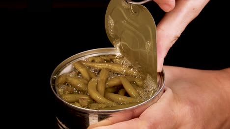 a hand opens a pull-tab can of green beans against a black background, revealing the beans in liquid. bright, even lighting and steady close-up camera work