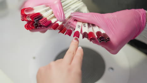 closeup view of customer pointing at preferred nail polish color from selection of vibrant red and burgundy shades held by nail technician wearing pink gloves during manicure consultation