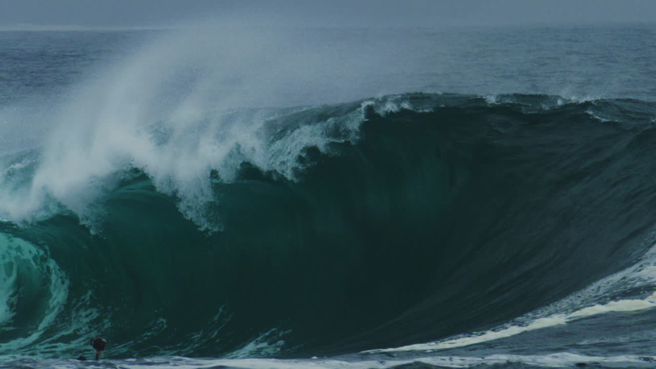 Telephoto view of dark green wave folding with force and throwing sea spray across frame in slow motion