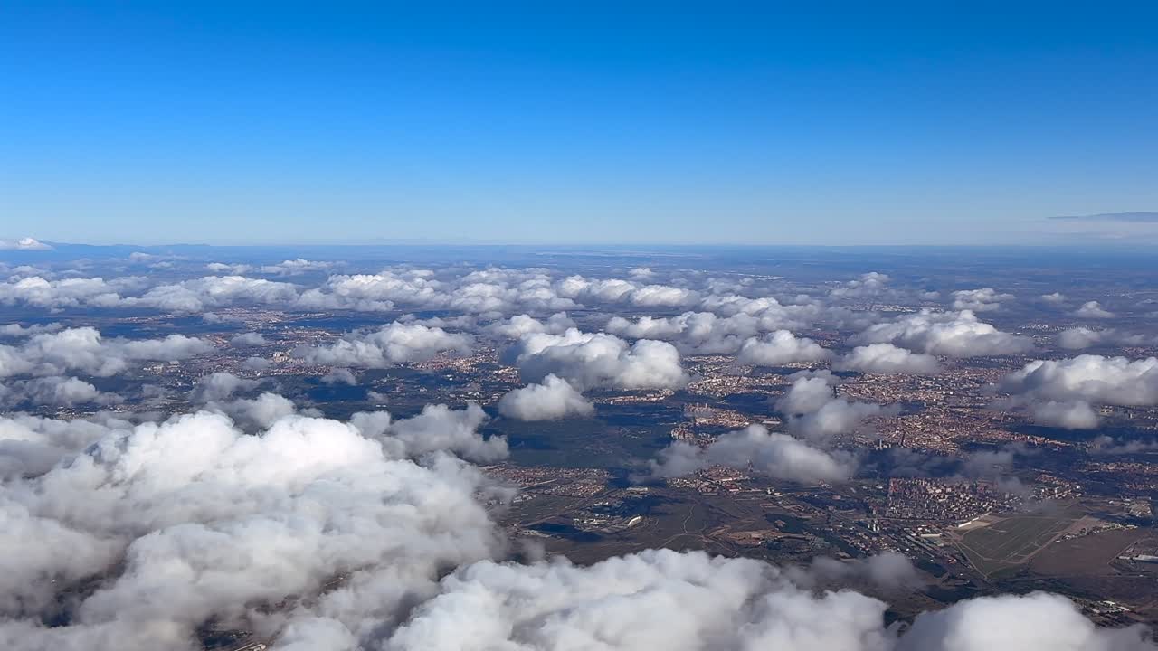 Et højtliggende luftfoto af Madrid og omegn taget fra et flys cockpit, med spredte skyer, der svæver over byen, på en solrig vintermorgen under en blå himmel.