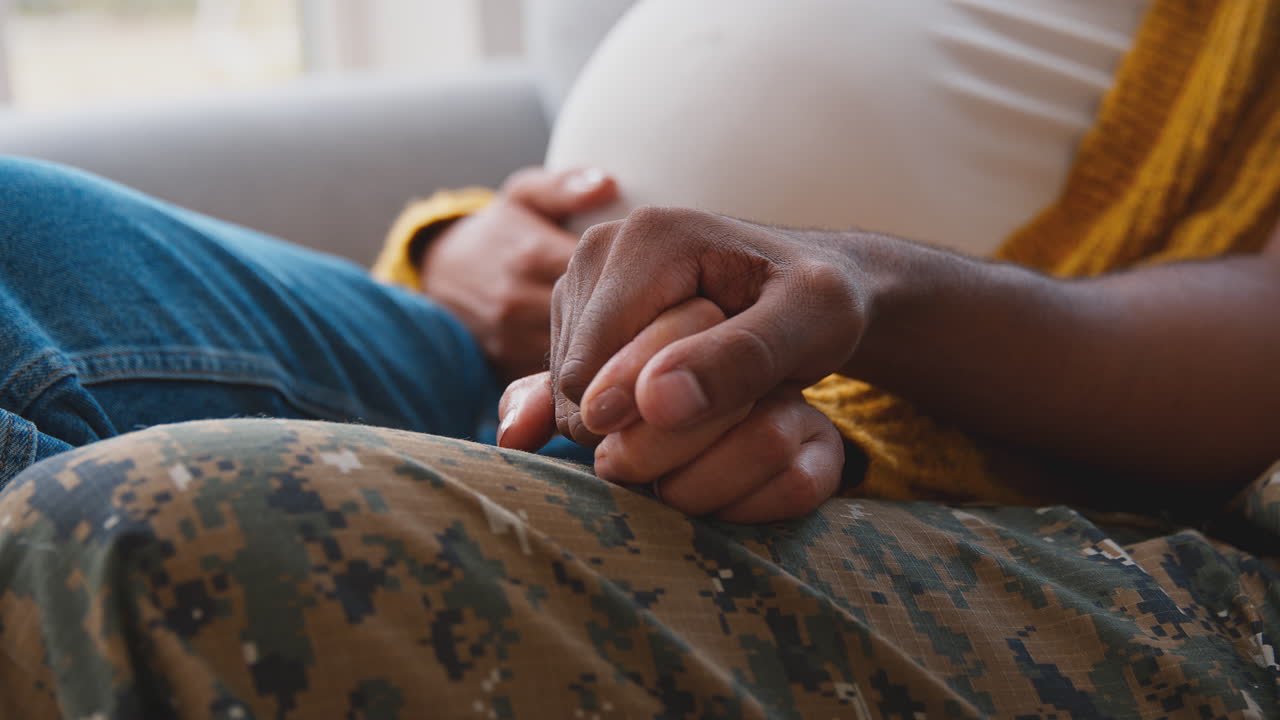 pareja embarazada con una mujer sentada al lado del marido del ejército en uniforme tomándose de la mano en casa de licencia