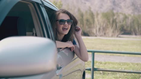 Una-Chica-Feliz-Con-Gafas-De-Sol-Se-Asoma-Fuera-Del-Coche-Contra-La-Pradera