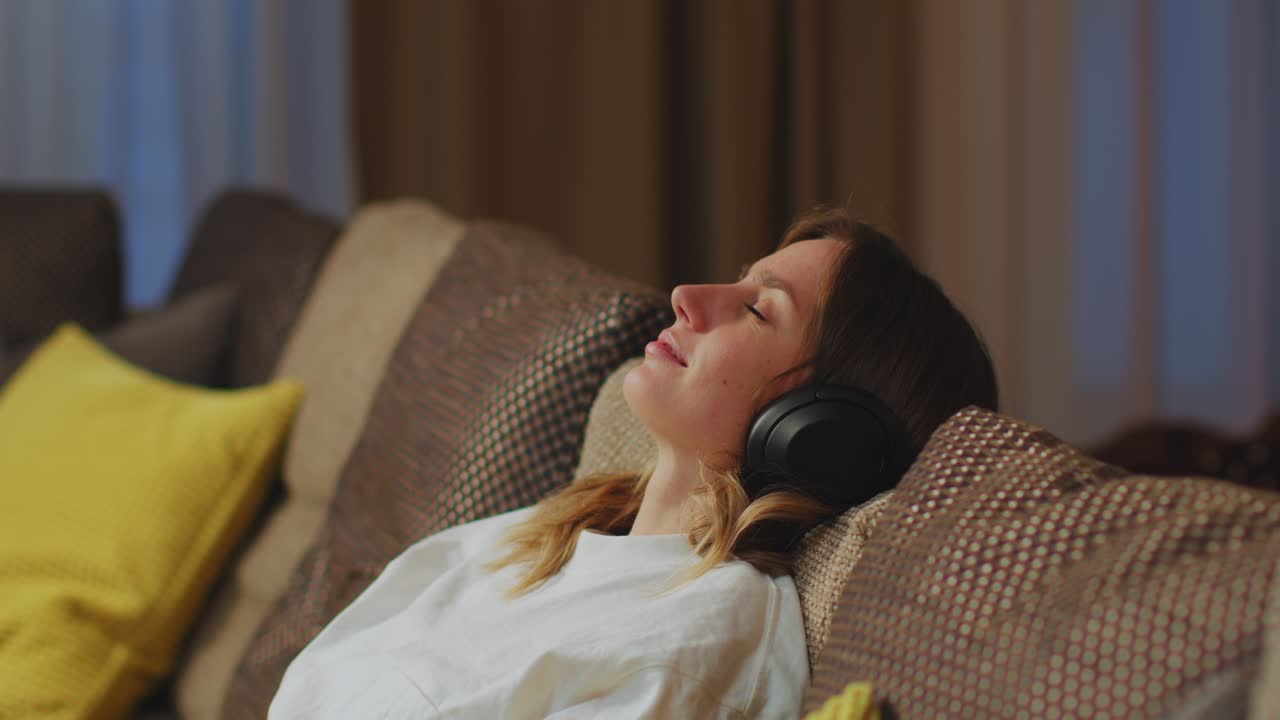 Calm young woman put on modern wireless headphones to relax on comfortable sofa, listening to music, stress-free concept