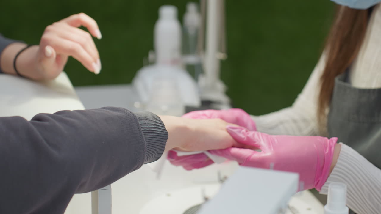 Close-up of nail technician wearing pink gloves cleaning client hand and nails during manicure session. Client rests hand on salon table while green wall and beauty products appear in background