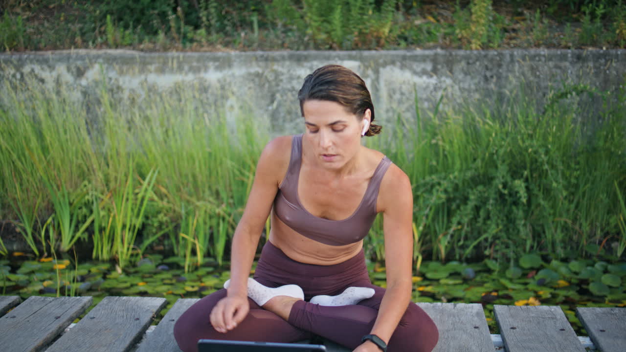 entrenador de yoga insertando auriculares en el banco del parque verde. mujer lección de tableta en línea