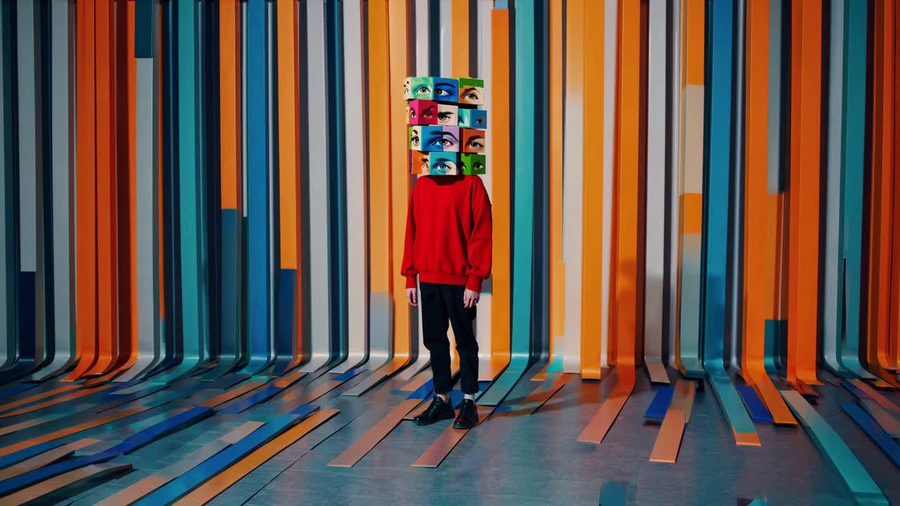 Person wearing red sweatshirt and black pants standing in vibrant room with colorful vertical elements, sporting cube shaped mask featuring playful eyes, creating dreamlike visual narrative
