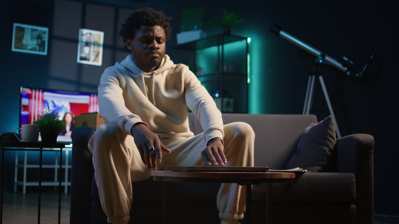 Ambitious black man learning a new language alone in his cozy living room, browsing internet for study platforms and explanations. Adult using language learning software online on laptop. Camera B.