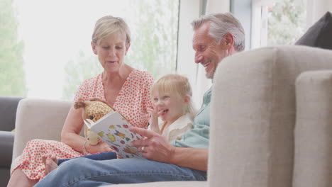 Großeltern-Sitzen-Mit-Enkelin-Zu-Hause-Auf-Dem-Sofa-Und-Lesen-Gemeinsam-Ein-Buch