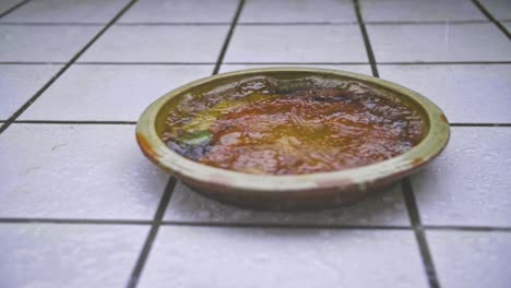 close-up pan of slow-motion rain flooding a brown flower clay jar with white tiles in background