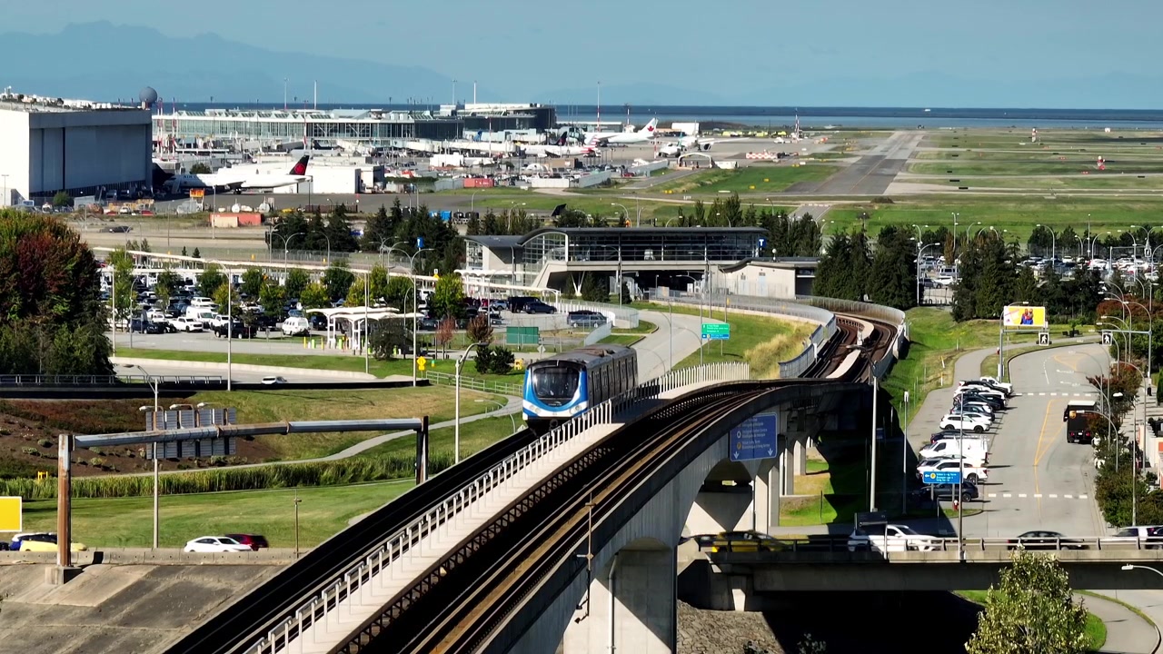 Um SkyTrain desliza por uma via elevada, com aviões e pistas de aterragem ao fundo perto de YVR na Colúmbia Britânica, Canadá - Plano Estático
