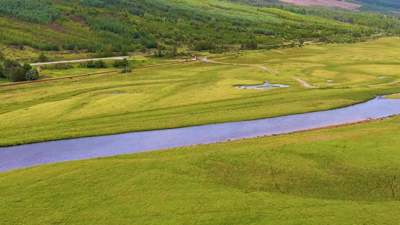 Drone glides above tranquil river, wooden bridge, green meadows, forest, and distant Scottish hills