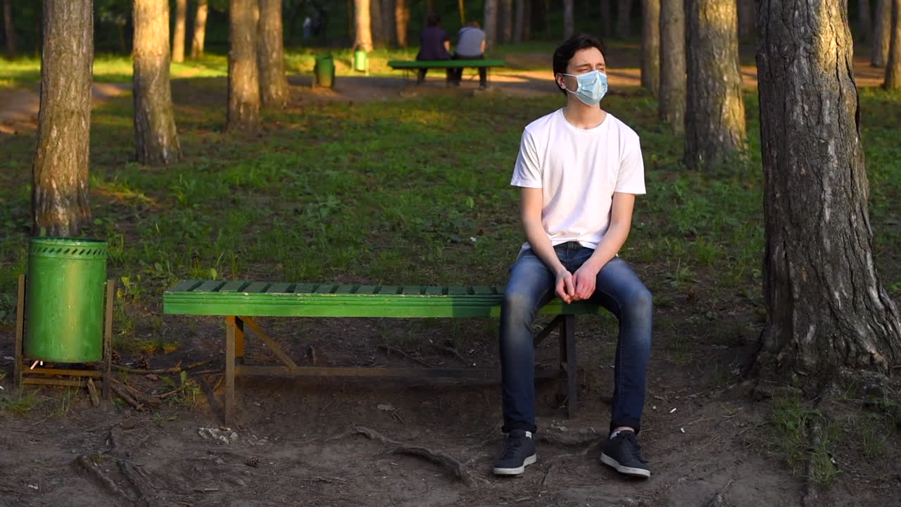 Lonely young man in white t-shirt and protective medical mask enjoying nature and looking around while resting on the bench in the park. Sunny day. Corona Virus idea