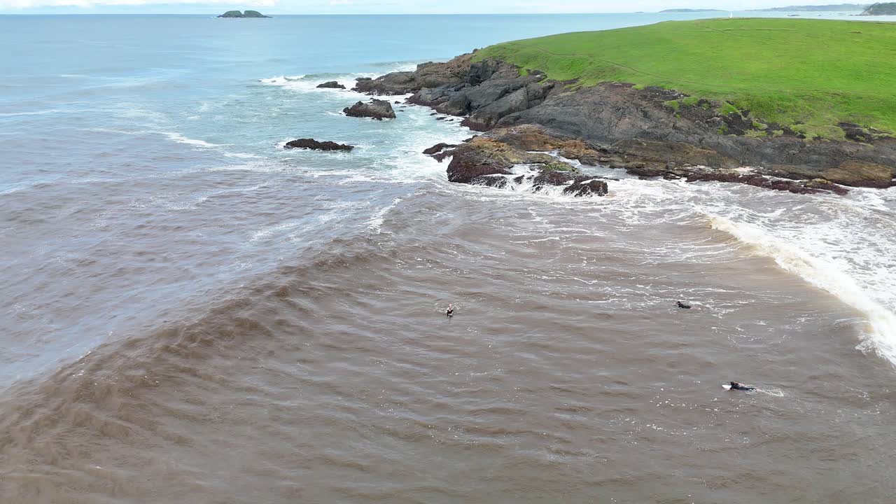 Aerial view of surfers catching brown river plume waves near rocky coastal headland, daylight