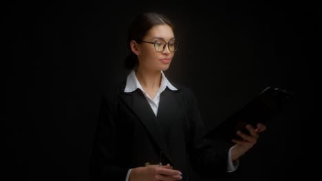 woman in glasses and business clothes signs a document attached to a tablet.