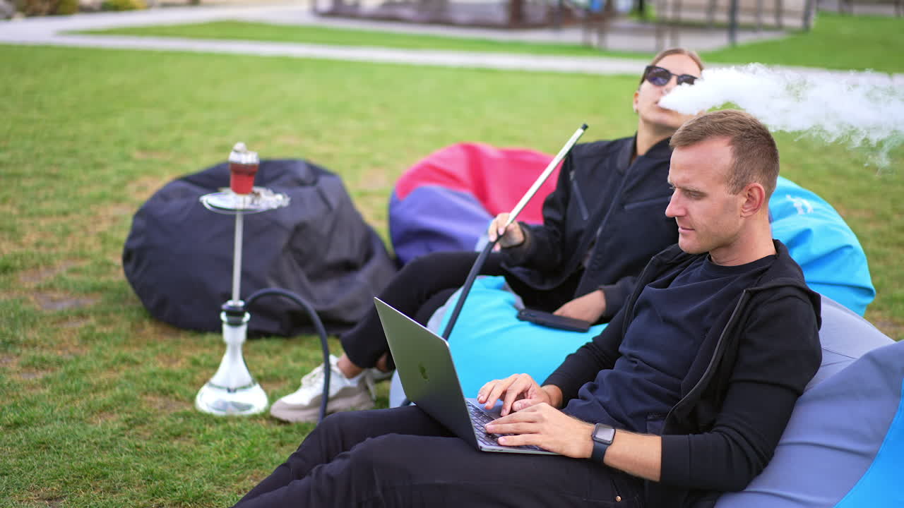 Modern couple spending time together outdoors. Man working on laptop and woman smoking hookah. Leisure time in bean bag chairs outdoors.