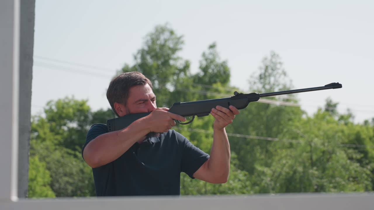 side view of man shooting rifle at pest in background near farm structure, rural greenery, focused expression, protective action to keep crops safe from birds or rodents