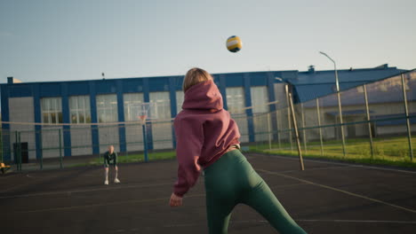 back view of woman attempting to serve volleyball but doing it incorrectly with two others in the background, one in green hoodie and another in blue hoodie, on volleyball court