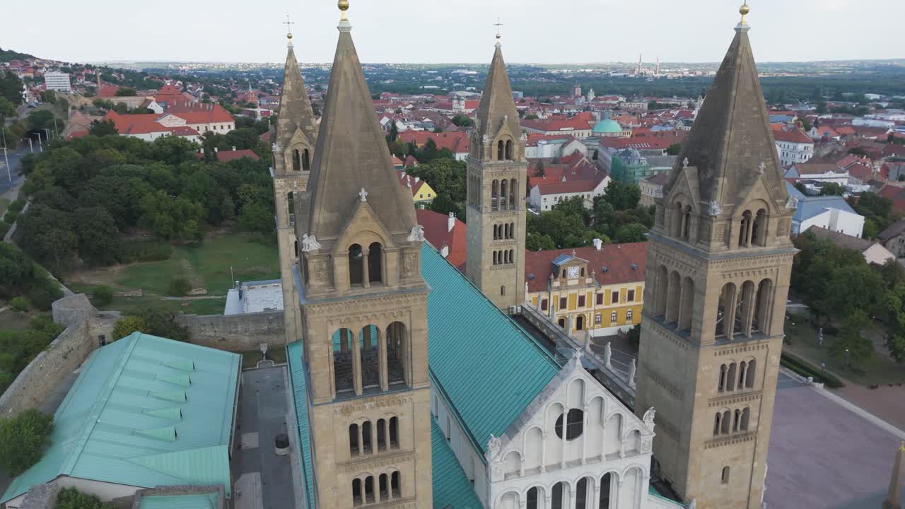 Drone orbiting Pécs Cathedral with birds flying, highlighting its twin towers, blue roofs, and historic old town backdrop