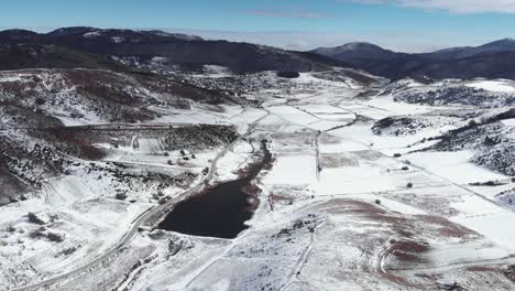 Aerial-birds-eye-view-snowy-mountain-fields-lake-peaks-at-distance-sunny-day