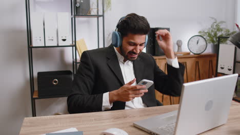 Indian-business-man-in-headphones-listening-favorite-disco-music,-working-on-laptop-at-home-office