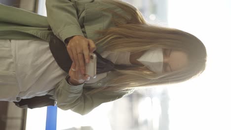 Young-relaxed-woman-in-mask-using-personal-cell-phone-for-chatting-with-friends-online-while-waiting-for-flight
