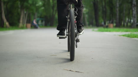 rear view of person in sneakers standing while riding bicycle on paved road, captured from low angle, in the background, there's a blurred view of another person sitting on a bench