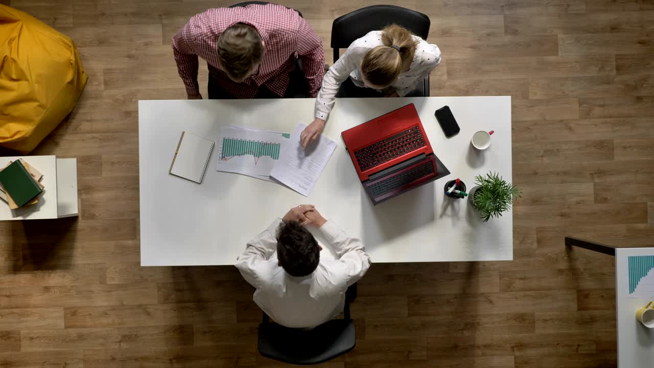 Team af tre unge forretningsfolk ankommer og diskuterer projekt, set ovenfra, siddende ved bord i moderne kontor.