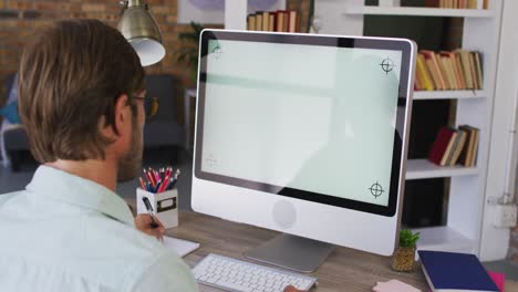 Caucasian-male-teacher-taking-notes-while-having-a-videocall-on-computer-in-the-classroom-at-school