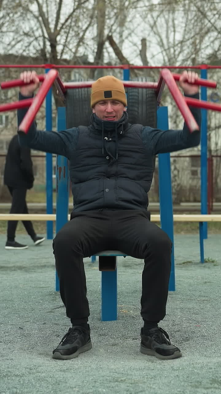 un entrenador con una gorra marrón y una chaqueta negra está trabajando en equipos al aire libre, levantando barras rojas sobre su cabeza, en el fondo, un niño camina, con coches que pasan en la distancia