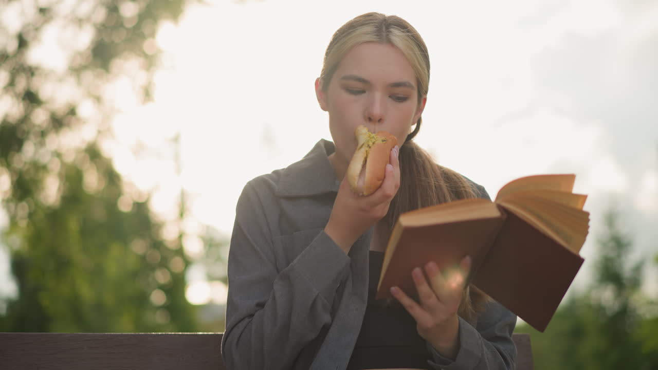 dama de camiseta gris comiendo bocadillo mientras lee un libro al aire libre, sentada en un banco del parque rodeada de árboles y vegetación con fondo borroso, la luz solar natural ilumina suavemente la escena