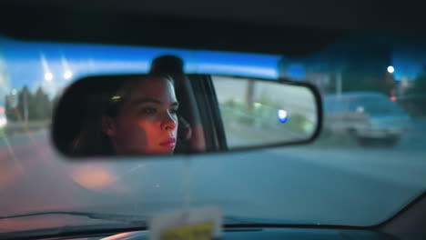 la mujer se refleja en el espejo retrovisor mientras conduce su coche por la noche, con coches estacionados y edificios cercanos visibles en el fondo bajo una iluminación tenue en una carretera urbana con una marca amarilla