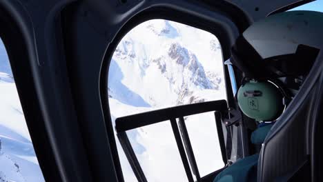 helicopter pilot looking out the side window at bright white snowy rocky mountains