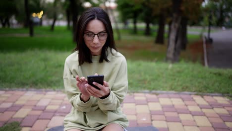 una chica morena con un suéter verde y gafas se sienta en una patineta en el parque y envía mensajes por teléfono. paseo por la noche, ocio