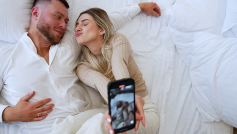 happy caucasian couple lie on the bed taking selfie. people in love kiss lovingly. top view.