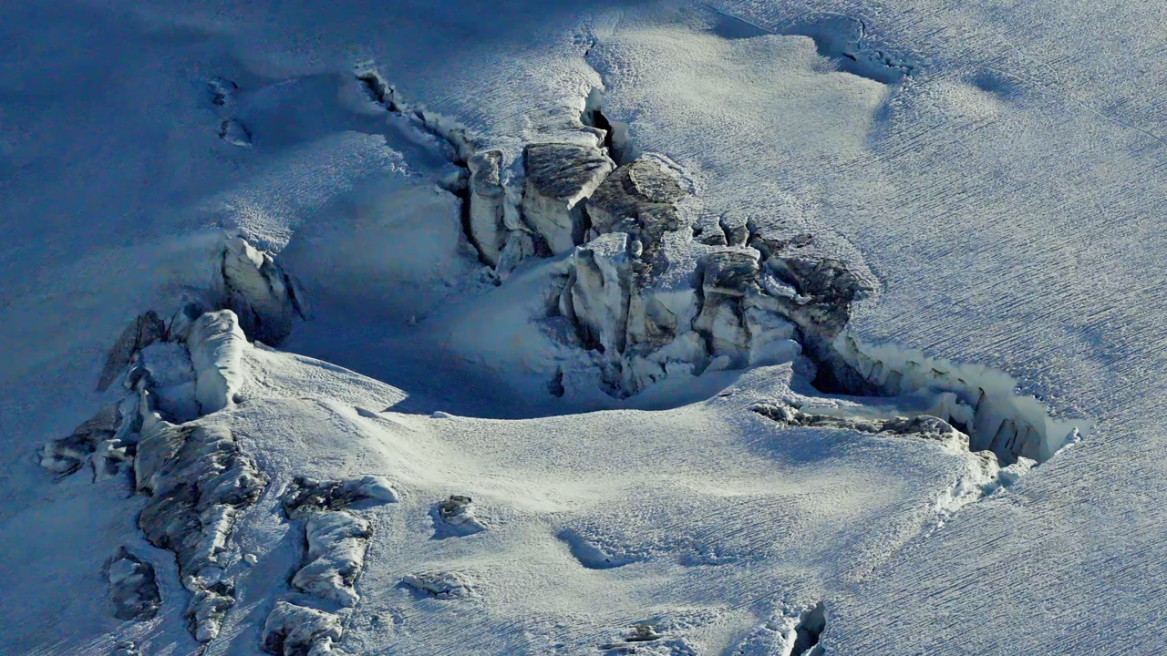 An aerial close-up of a deep glacial crevasse and ice fracture in the Chamonix Alps of France, showing the textured blue ice and snowy terrain of the Mont Blanc massif on a sunny winter day