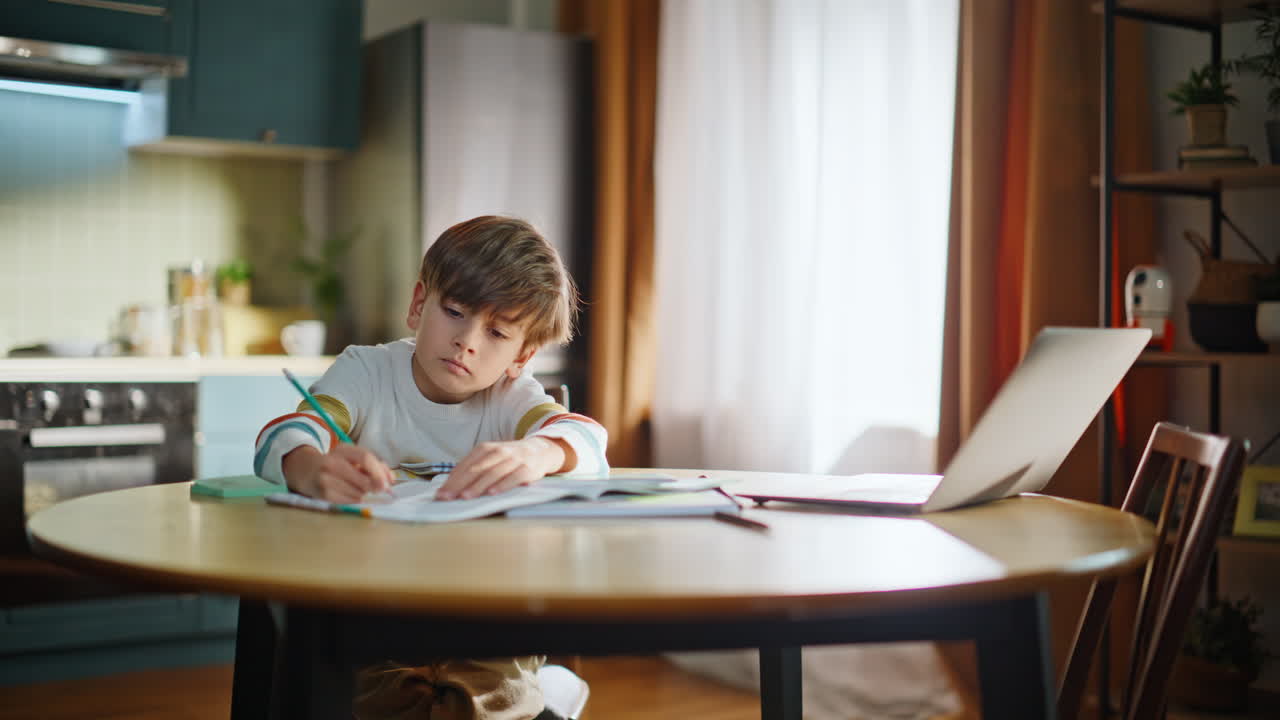 Pensive schoolboy pondering homework writing copybook sitting kitchen desk alone