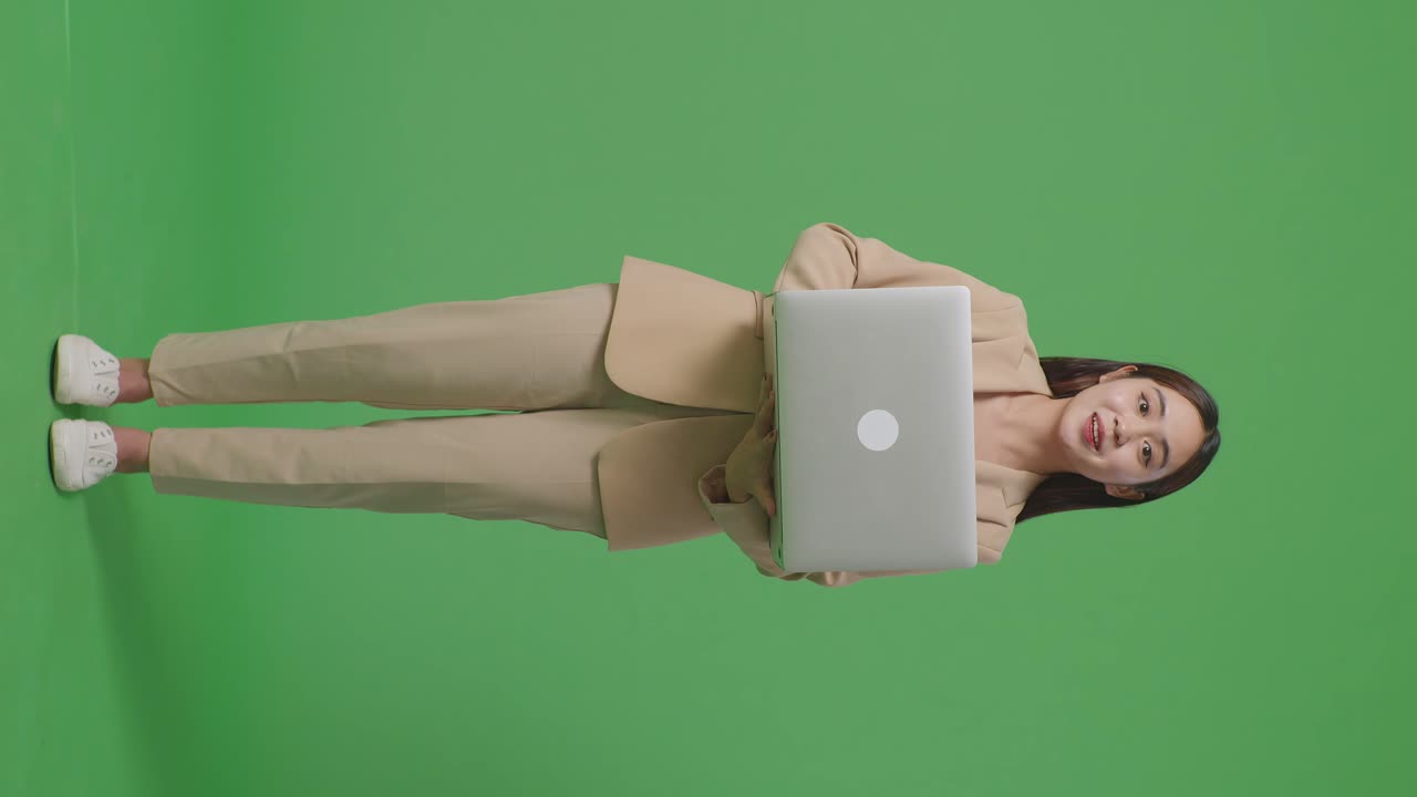 cuerpo lleno de mujer de negocios asiática usando computadora portátil y sonriendo a la cámara en el fondo de la pantalla verde en el estudio