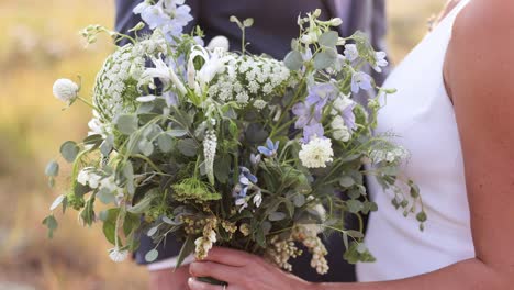 close up of bride in her wedding dress holding large wildflower wedding bouquet in her hands in wedding dress outdoors with groom 1080p 60fps