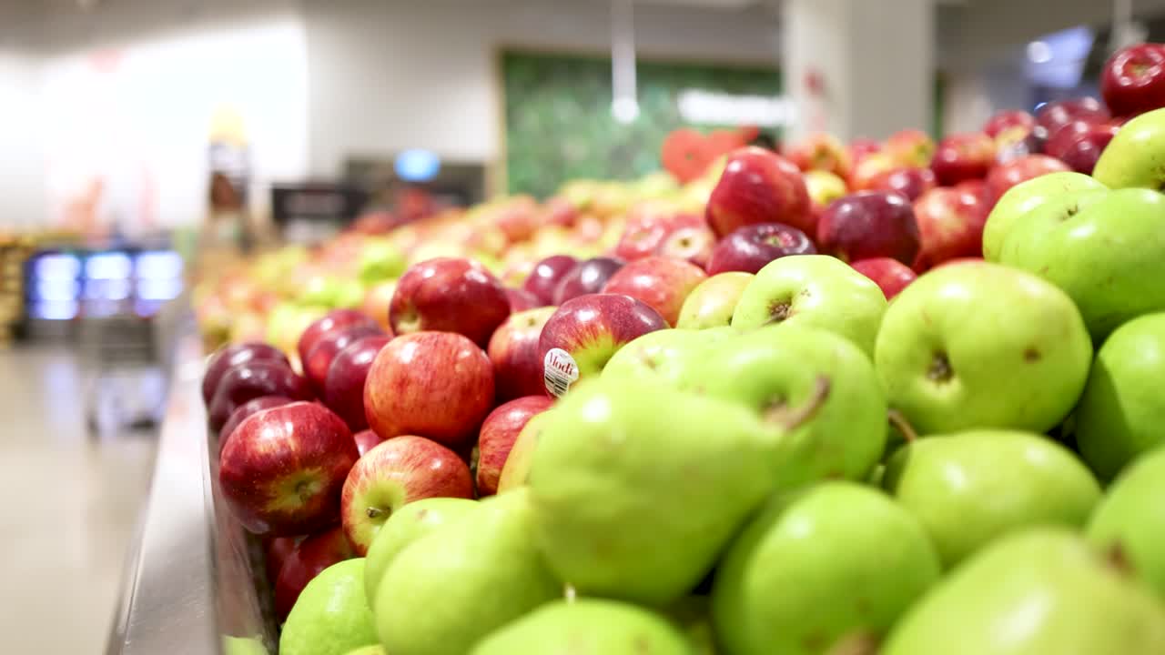 Brightly lit supermarket aisle with red and green apples. Shoppers move in the background, creating a lively atmosphere
