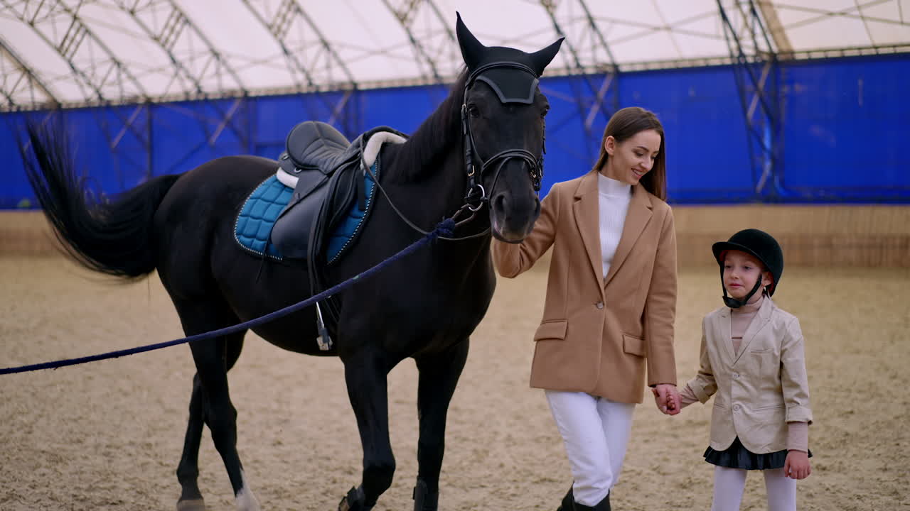 Lady and her little daughter attending horse riding lessons at drill-hall. Woman is holding a beautiful black horse by the bridle.